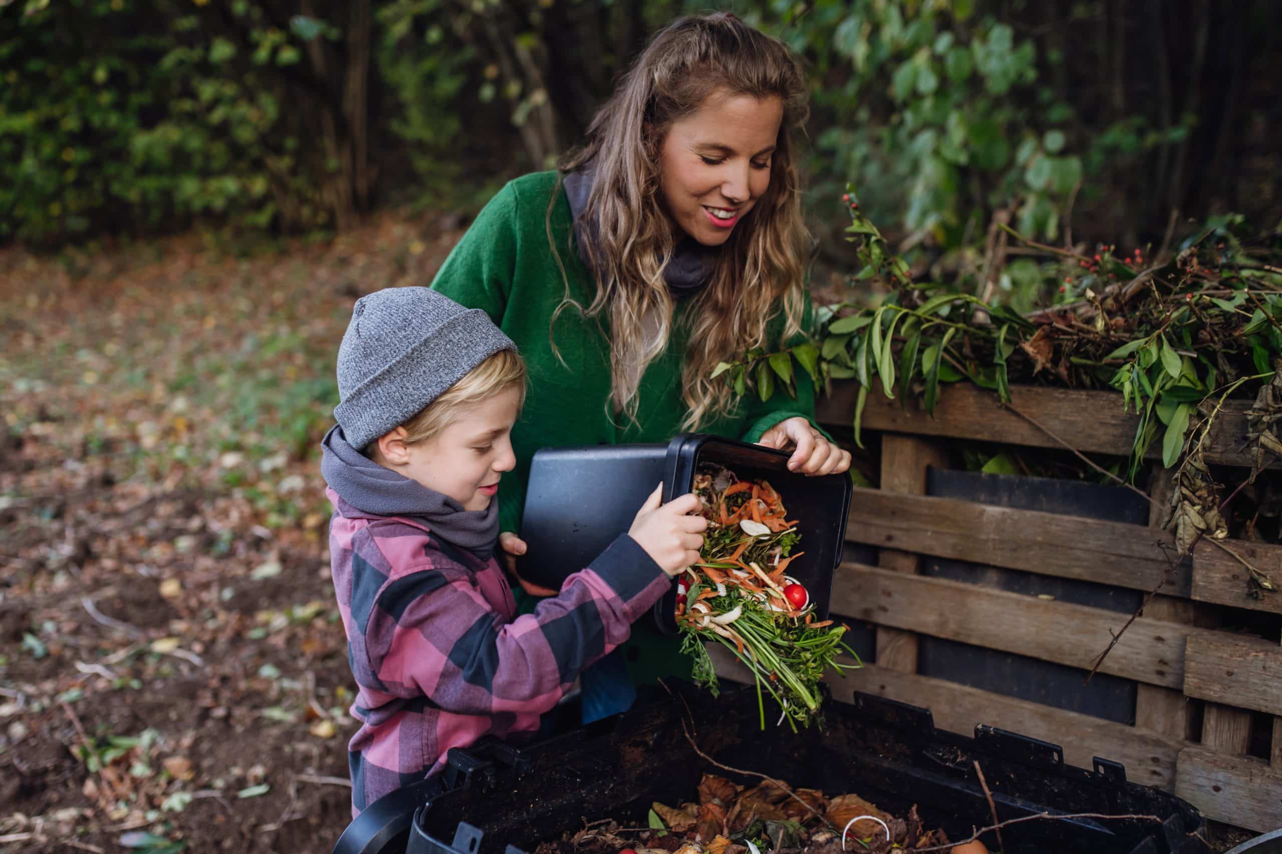 Boy helping mother to put kitchen waste, peel and leftover vegetables scraps into composter in the garden. Concept of composting kitchen biodegradable waste.